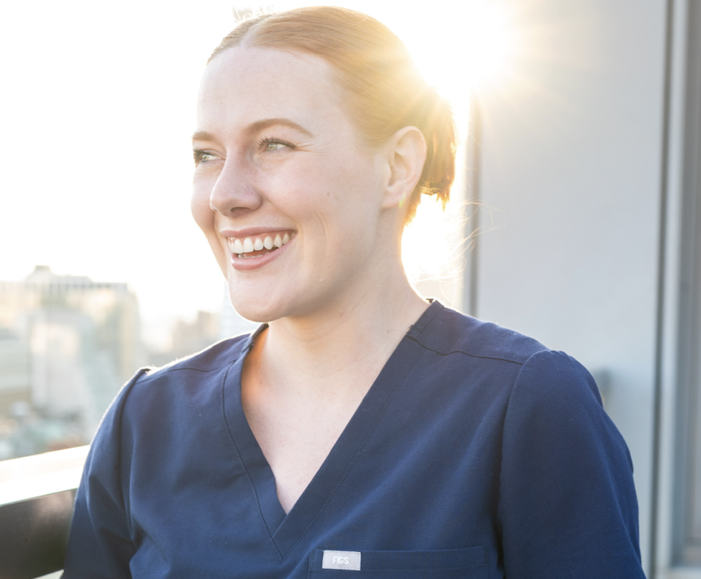 Smiling nurse in blue scrubs standing near a bright window.